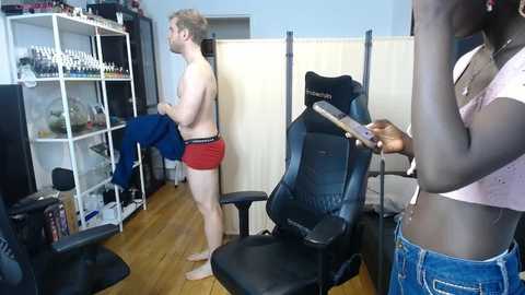 Media: Video of a room with a man in red shorts and a woman in a pink top, standing by a black gaming chair and wooden floor.