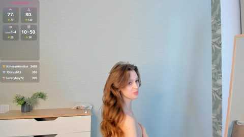 Media: A video of a nude, light-skinned woman with long, wavy brown hair, standing indoors near a white wall with a digital thermometer.