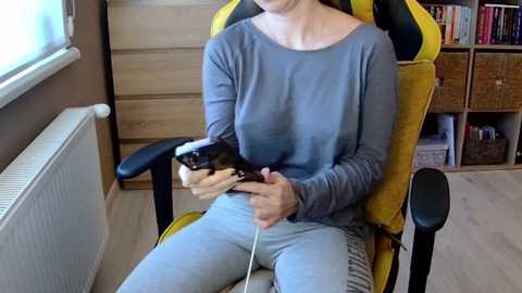 Media: Video of a light-skinned woman with medium-length brown hair, seated in a mustard-yellow gaming chair, wearing a grey long-sleeve top and light blue leggings, holding a black PlayStation controller, indoors with wooden paneling and bookshelves in the background.