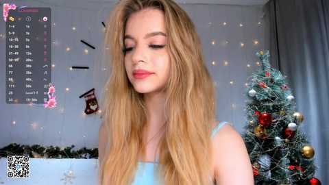 Media: Video of a blonde woman with long, wavy hair, wearing a light blue tank top, standing in a festive room with a decorated Christmas tree, calendar, and holiday decor.