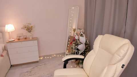 Media: Video of a cozy, pastel-themed bedroom with a white leather armchair, white dresser, and a tall, mirrored floor lamp with fairy lights.