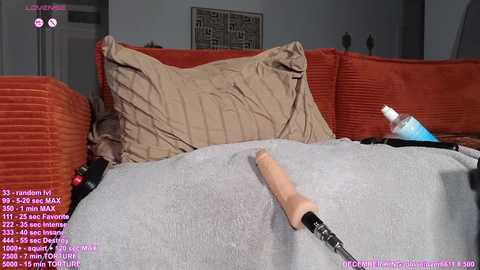 Media: Video of a cozy living room featuring an orange couch with a beige blanket, a beige pillow, and a nude-colored sex toy. The room has gray walls with a framed art piece.