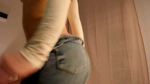 Media: A video of a person from the waist down, wearing light-colored, long-sleeved clothing and light blue jeans, standing in a room with wooden walls.