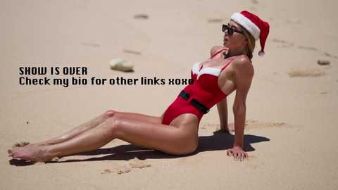 Media: A video of a slender, fair-skinned woman in a red one-piece swimsuit and Santa hat lounging on sandy beach, legs stretched out, with text overlay.