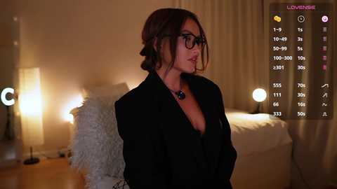 Media: Video of a brunette woman in glasses, wearing a black blazer, in a cozy bedroom with warm lighting, white bedding, and a fluffy pillow, with a \"Controlling\" overlay.