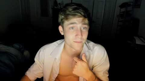 Media: Video of a young, shirtless white male with short brown hair, looking into the camera with a serious expression, in a dimly lit room with a bed, wardrobe, and dark background.