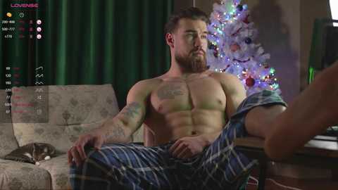 Media: Video of a muscular, bearded man with tattoos, shirtless, lounging on a couch in plaid pajamas, next to a decorated Christmas tree. Background includes a green curtain and a beige sofa.