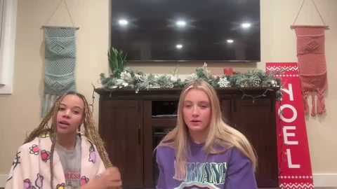 Media: Video of two young girls with braided hair, wearing holiday-themed sweaters, in a cozy living room with a flat-screen TV, Christmas decorations, and festive wall hangings.