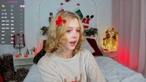 Media: A video of a young blonde woman with wavy hair, wearing a festive sweater, sitting on a bed in a cozy, Christmas-decorated room.