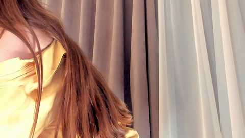 Media: A video showing a woman's long, straight brown hair cascading over her shoulders, partially obscured by a mustard-yellow off-the-shoulder blouse. The background features light gray and white sheer curtains, creating a soft, elegant ambiance.