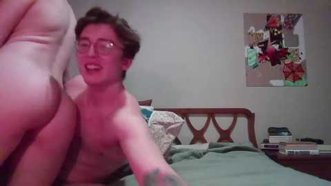 Media: Video of a young, shirtless, fair-skinned man with glasses and short hair, smiling while standing beside a woman in a white dress, in a messy bedroom with a cluttered nightstand and colorful pinboard.