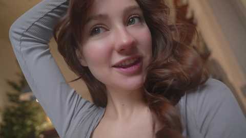 Media: Video of a smiling, fair-skinned woman with long, wavy brown hair, wearing a gray cardigan, taken indoors, with blurred background.