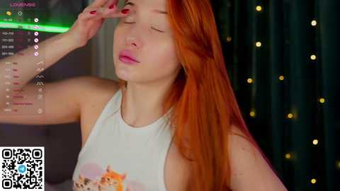 Media: Video of a fair-skinned woman with long, fiery red hair wearing a white tank top, wiping her forehead, in a dimly lit room with string lights and a green screen in the background.