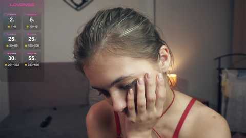 Media: Video of a young Caucasian woman with light brown hair in a ponytail, wearing a red tank top, covering her face with her hands in a distressed pose. Background includes a dimly lit room with a bed, blurred out.