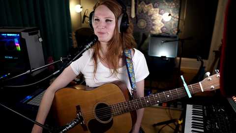 Media: Video of a young woman with long red hair, wearing headphones and a white t-shirt, playing an acoustic guitar in a dimly lit, colorful, bohemian room with instruments and stage lighting.