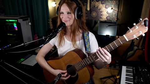 Media: Video of a smiling, long-haired woman with headphones, playing a ukulele while singing into a microphone in a dimly-lit, bohemian-themed studio with psychedelic tapestry and music equipment.