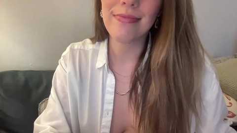 Media: Video of a smiling woman with long brown hair, wearing a white button-up shirt open at the collar, revealing cleavage. She's seated on a couch with a beige cushion, in a cozy indoor setting.