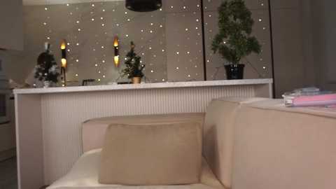 Media: Video of a modern, minimalist kitchen with beige walls, white cabinets, and a white marble countertop. Features a beige couch, a potted plant, and decorative string lights.