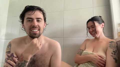 Media: A video of a man with wet, short black hair, smiling, and a woman with wet dark hair, covered in a towel, in a beige-tiled bathroom.