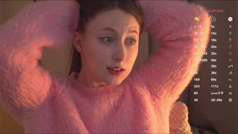 Video of a young woman with fair skin and brown hair, wearing a pink fuzzy sweater, smiling while adjusting her hair. Background includes a computer screen displaying a chat interface.