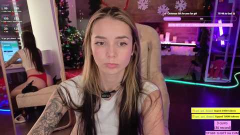 Media: Video of a young woman with long, straight blonde hair, wearing a white top and a black choker, sitting in a modern room with a Christmas tree and glowing lights.