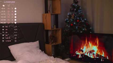 Video of a cozy bedroom with a dark gray headboard, white bedding, and a lit fireplace in the background. Adjacent is a wooden shelf with a decorated Christmas tree.