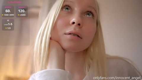 Media: Video of a young, fair-skinned blonde woman with blue eyes, resting her chin on her hand. The background is blurred, featuring a light-colored wall. The image is accompanied by a screen overlay with streaming metrics.