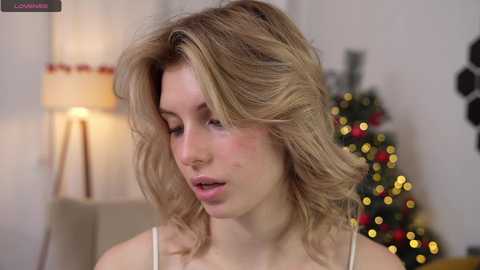 Media: Video of a blonde woman with wavy hair, light skin, wearing a white camisole, indoors, near a lit Christmas tree, blurred background.