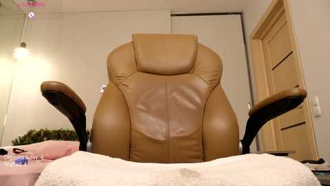 Media: Video of a tan leather massage chair with adjustable armrests, positioned in a softly lit bedroom with white walls, a pink blanket, and a closed beige door in the background.