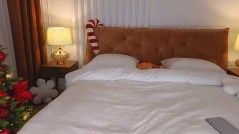 Media: Video of a cozy, holiday-themed bedroom with a tufted, brown headboard, white linens, a festive, red-decorated Christmas tree, and a glowing, gold lamp on a wooden nightstand.