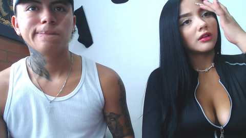 Media: A video of a young, light-skinned Hispanic woman with long black hair and large breasts, wearing a low-cut black top, and a young, light-skinned Hispanic man with a sleeve tattoo, wearing a white tank top.