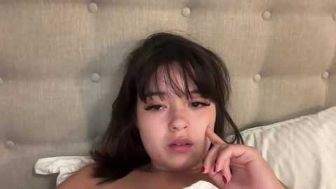 Media: A video of a young Asian woman with shoulder-length dark hair, lying on a beige, tufted headboard, with her face partially covered by a white pillow.