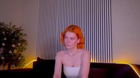 Media: Video of a fair-skinned woman with orange hair, wearing a strapless white top, seated on a dark couch in a dimly lit room with vertical blinds and a lit Christmas tree in the background.