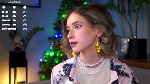 Video of a young woman with wavy blonde hair, wearing a pink floral blouse, pearl necklace, and yellow earrings, standing in a festive room with a lit Christmas tree and potted plants.