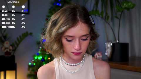 Video of a young, fair-skinned woman with wavy blonde hair, wearing a pearl necklace, sitting indoors beside a decorated Christmas tree.