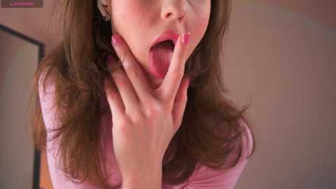 Media: Video of a young woman with long brown hair, wearing a pink top, playfully sticking out her tongue and touching her face with painted nails.