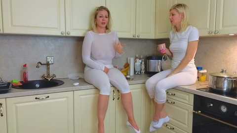 Media: Video of two blonde women in white pajamas sitting on a kitchen counter, one with a hand on her chest, the other holding a pink phone, in a modern, cream-colored kitchen.