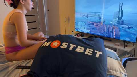 Media: A young woman with light skin, wearing a pink bra and panties, plays a video game on a bed. A large dark blue pillow with \"IMOSTBEET\" text covers her lap. A TV screen shows an underwater scene in a submarine.