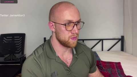 Video of a bald, bearded man with glasses, wearing a green polo shirt, sitting on a bed with a red plaid blanket, in a simple bedroom with a black chair and white walls.