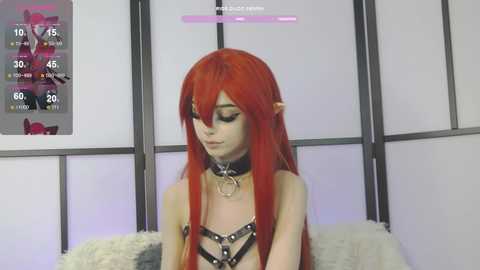 Media: Video of a young woman with long, bright red hair, wearing a black leather harness and choker, sitting on a white furry cushion. Background includes a pink and purple digital clock and a wall with black and white panels.