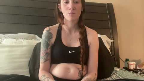 Media: Video of a tattooed woman with long brown hair, wearing a black sports bra, sitting on a bed in a bedroom.