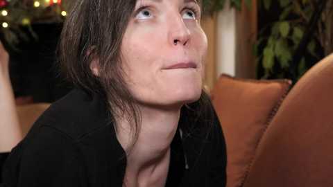 Media: Video of a woman with shoulder-length brown hair, wearing a black shirt, looking contemplative with slightly parted lips, sitting on a tan leather couch, blurred background with Christmas decorations.