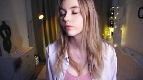 Media: Video of a young Caucasian woman with long, straight blonde hair, wearing a pink tank top and white blouse, standing in a dimly lit bedroom with beige walls, a bed, and a desk in the background.