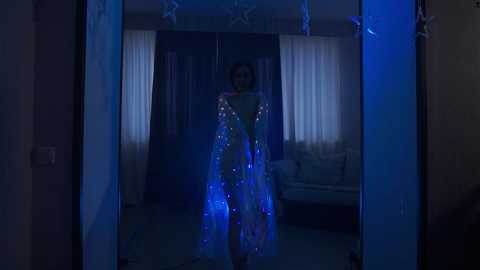 Media: Video of a dimly lit room with a person wearing a translucent, blue, glittery dress, standing in front of a window with sheer curtains. The room has a minimalist decor with neutral tones and soft lighting.