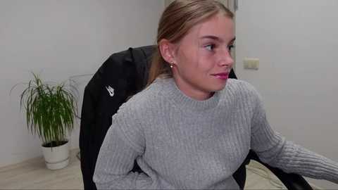 Media: Video of a young, light-skinned woman with blonde hair, wearing a grey ribbed sweater, seated on a black office chair in a minimalist, white-walled room with a potted plant and a white wall outlet in the background.