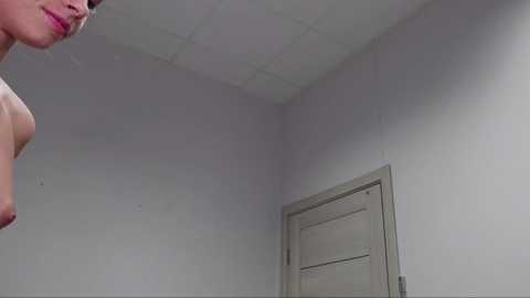 Media: Video of a naked woman from the shoulders up, in a plain white room with a closed door and ceiling tiles.