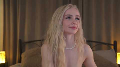 Media: Video of a fair-skinned, blonde woman with long, wavy hair, wearing a pearl necklace, seated on a bed with beige pillows, in a dimly lit room with brown curtains and two bedside lamps.