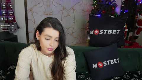 Media: Video of a young woman with long, dark hair, wearing a beige sweater, sitting on a couch with a dark patterned blanket. Behind her, a decorated Christmas tree and a black cushion with \"MOSTBET\" logo.