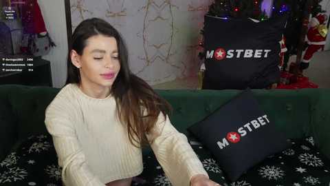 Media: Video of a young woman with long, straight brown hair wearing a cream sweater, seated on a green couch with black pillow featuring \"MastBet\" logo, in a festive, Christmas-decorated room.