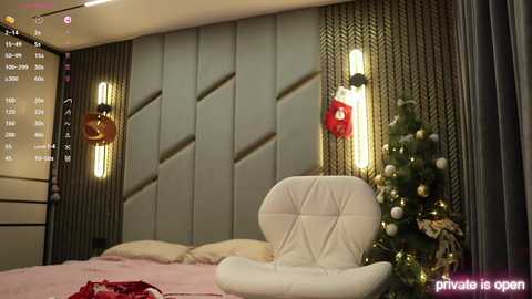 Media: A video of a modern, minimalist bedroom with a beige padded headboard, a white leather armchair, a lit Christmas tree, and wall-mounted sconces.